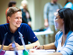 A male student smiling working with a femail student on laptops in the classroom