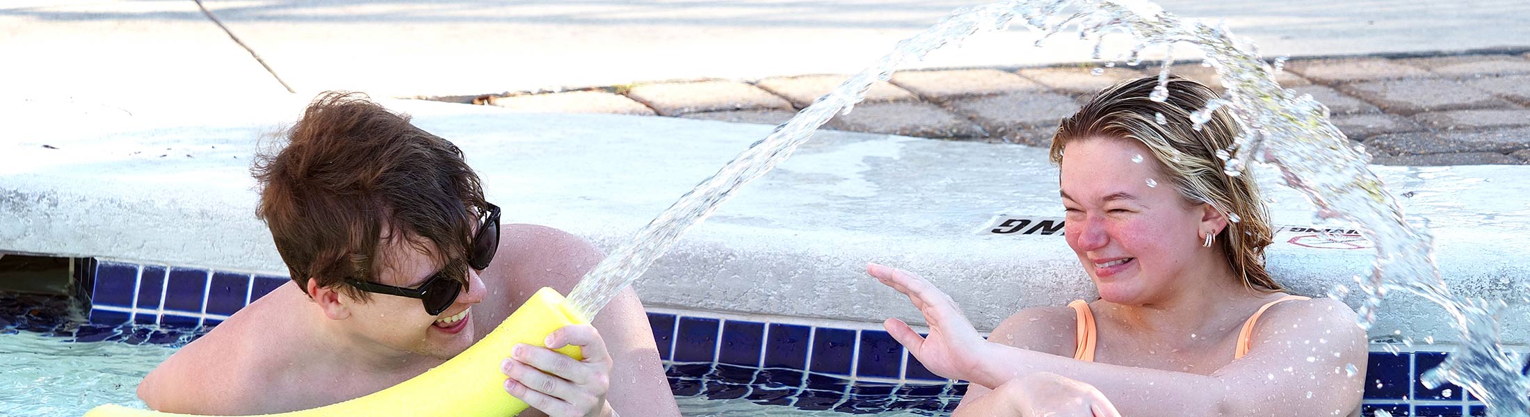 Students playing with pool noodles in campus rec outdoor pool.